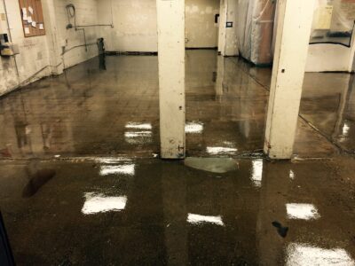 An industrial space with wet concrete floors showing reflections of overhead lights. Two prominent, white-painted pillars stand in the center. The walls appear aged with peeling paint and notice boards hanging. Puddles on the floor indicate recent cleaning or a leak. The area behind the pillars is partitioned off with plastic sheeting, suggesting ongoing maintenance or construction work.