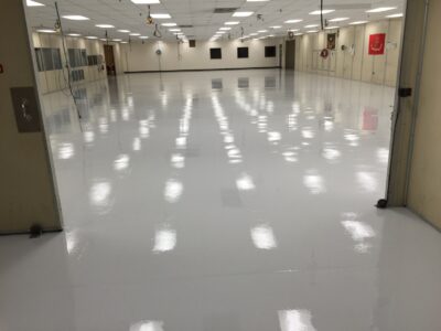 ALT Text: "A vast, empty industrial space with a high-gloss white epoxy floor reflecting the fluorescent lights overhead, creating a pattern of soft glows across the surface. The room has multiple bays with closed overhead doors on the right side, each adorned with various national flags. Power outlets and emergency equipment like a fire hose and fire extinguishers are strategically placed along the walls. The spacious and clean environment suggests a setting prepared for activities requiring sterile or controlled conditions.