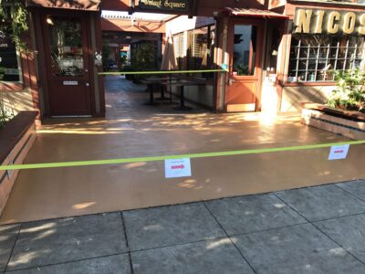 An outdoor dining area cordoned off with yellow caution tape and 'Do Not Enter' signs posted on it, indicating a restricted or closed-off space. The area features a smooth tan-colored floor that transitions to cobblestone pavement. Two restaurant entrances are visible, one with the sign 'Walnut Square' and the other 'NICOS', both showcasing dark wooden doors and warm lighting. Greenery in planters adds to the ambiance. The setting sun casts a soft glow and shadows, suggesting the photo was taken in the late afternoon or early evening.