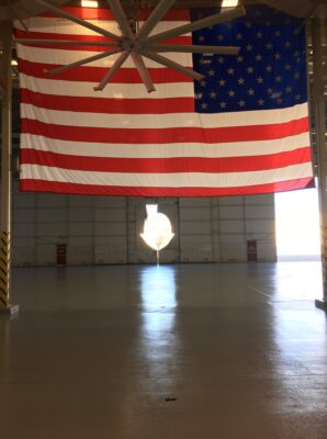 A large American flag hangs prominently on the back wall of a spacious industrial hanger, with sunlight streaming through the open door behind it, creating a bright silhouette effect. The hanger features a high ceiling with a large metallic fan, which is partially obscured by the flag. The floor is polished concrete, reflecting the natural light. Yellow and black safety stripes are visible near the hanger entrance, with the interior otherwise unoccupied, providing a sense of scale and grandeur to the setting.