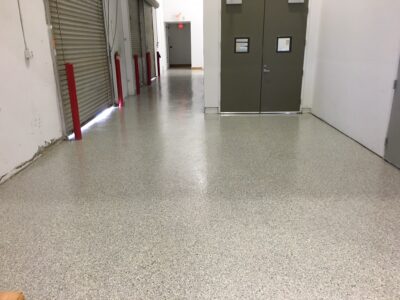 A commercial building corridor with an epoxy-coated floor, showing a blend of grey and speckled colors. On the left, there are several closed metallic rolling shutters, each guarded by a red bollard. The white walls are marked from wear, and electrical outlets are visible. Further down the corridor is a set of closed double doors in dark grey with rectangular windows and nameplates. Fluorescent lighting reflects off the shiny floor surface, and the corridor leads towards an exit sign in the background, suggesting depth and passage.