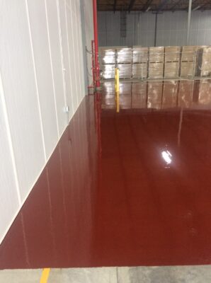 Interior of a warehouse showing a glossy, red-painted concrete floor with reflections visible on the surface. The floor meets a white wall on the left and pallets stacked with cardboard boxes are visible in the background. Fire safety equipment, including a red fire hose reel, is mounted on the wall, and a yellow safety bollard stands in the foreground.