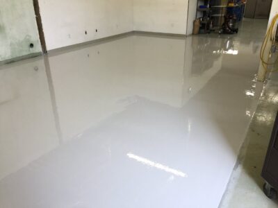 A newly renovated space with a fresh, glossy white epoxy floor that reflects the surroundings. On the left, an unfinished wall corner shows the raw concrete meeting the smooth flooring. Electrical outlets are spaced along the wall, ready for use. On the right, cleaning equipment and a yellow hose are stationed near a utility cart, suggesting ongoing work or cleaning. The room is brightly lit, enhancing the clean and open feeling of the space.
