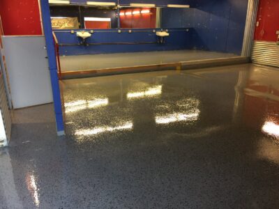 An indoor car wash bay with wet, glossy black and grey speckled epoxy flooring reflecting the bright overhead lights. The walls are painted blue with red and white accents, and two sinks are mounted against the back mirror reflecting the space. A metallic garage door is partially open to the right, and yellow safety lines demarcate the washing area. The environment suggests the bay is ready for vehicle cleaning operations.