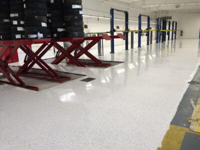 Inside an automotive service area with a clean white epoxy floor speckled with grey and black dots. To the left, a stack of black car tires is stored on a red multi-tier rack. The floor reflects the lighting and colors of the room, enhancing the sense of cleanliness and order. Multiple blue car lifts with yellow safety bars stand in a row towards the right, ready for vehicle maintenance. The floor is marked with yellow and blue lines, indicating work zones and paths for safety and organization.