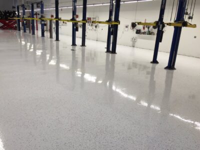 A view of a vehicle maintenance workshop with a shiny white epoxy floor that is speckled with black dots. The floor reflects the blue vehicle lifts and the bright overhead lights. Each lift is equipped with yellow safety locks. The back wall is equipped with various automotive tools and hoses. This clean and well-organized workspace is indicative of professional standards and attention to detail in the automotive industry.