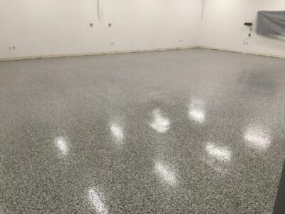 Spacious room with speckled epoxy flooring in varying shades of grey. The floor's reflective surface casts diffused reflections of the fluorescent lights overhead. White walls with electrical outlets and a covered section on the right suggest a space in transition or under preparation. The absence of furniture or other items provides a clean, open atmosphere suitable for industrial or commercial use.
