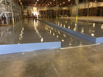 Industrial warehouse interior with freshly applied epoxy floor coating reflecting overhead lights. A long, elevated blue ramp divides the space, leading to an area with pallets of stocked goods. The vast, glossy floor extends throughout the facility, indicating a clean and maintained work environment.