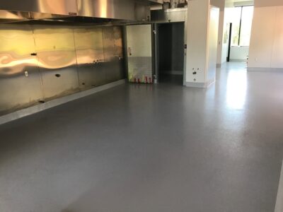 Wide-angle view of a spacious commercial kitchen with a smooth, light grey epoxy-coated floor. Reflective surfaces of stainless steel appliances and wall panels catch the natural light from the windows, creating a luminous environment. Visible is a large stainless steel ventilation hood, a diamond plate corner guard to the right, and fire extinguishers lined up on the left, indicating a focus on functionality and safety in the design.