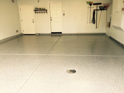 A clean and spacious garage interior with a freshly coated epoxy floor in a light grey shade, reflecting subtle specks of color. On the left wall, there's a small black shelf holding green gardening shoes and hooks beneath with hanging keys. To the right, a selection of garden tools, including rakes and brooms, is neatly organized against the wall. A white door with a window and a standard door is at the back, with a switch and an electrical outlet visible beside it. The floor features a central circular drain.