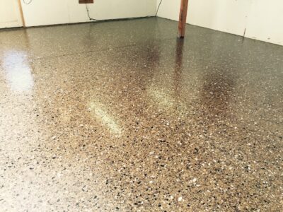 A room with a shiny, polished concrete floor scattered with multi-colored flakes, creating a terrazzo-like effect. Natural light reflects off the surface, highlighting the floor's texture. The walls are plain and white with an exposed wooden post on the right. Electrical wiring is partially visible along the back wall, suggesting an unfinished or utility space.