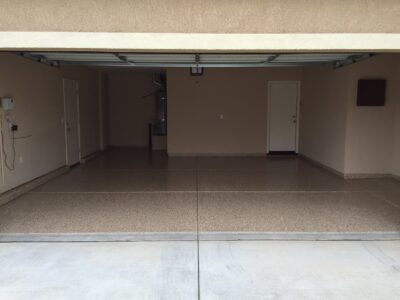 View from the driveway of an open, empty residential garage with a textured beige floor and walls. There is a white side door to the right, a smaller white door straight ahead, and a square window on the far right wall. Electrical outlets and a wall-mounted garage door opener are visible on the left side.