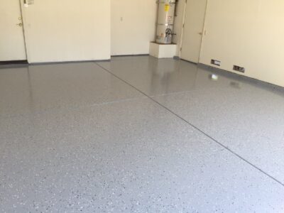 Interior view of a garage with a freshly applied epoxy floor coating. The floor is glossy and speckled with flakes of gray, white, and black, reflecting light from the surrounding area. The walls are white, and a water heater is installed in the corner. There are two vent covers on the wall near the floor.