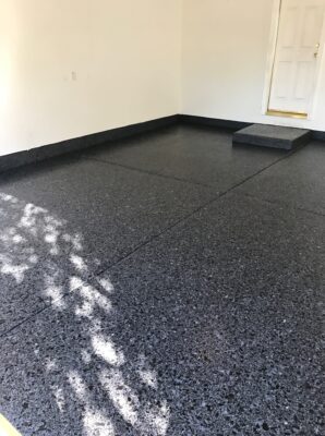 A corner of a room with a dark gray speckled epoxy floor featuring a raised platform step, natural light casting shadows on the floor, and a closed white door in the background.