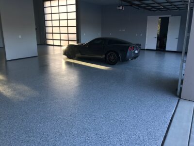 A dark gray sports car parked on a light gray speckled epoxy garage floor, with sunlight filtering through the partially open garage door.