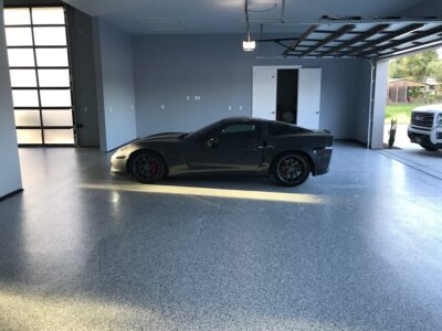 A dark gray sports car parked on a light gray speckled epoxy garage floor, with sunlight filtering through the partially open garage door.