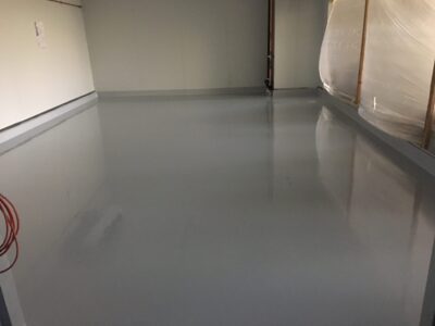 A newly coated light grey epoxy floor in an industrial space with subtle reflections, bordered by white walls and visible ductwork on the ceiling.