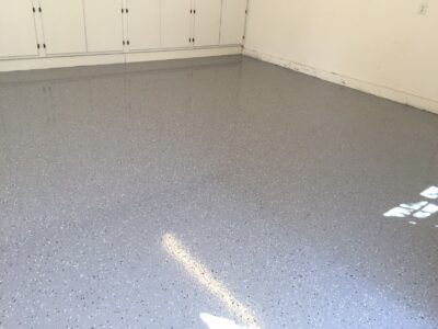 Interior view of a clean garage with a freshly applied grey, speckled epoxy floor coating. The smooth, shiny surface reflects sunlight from a window, casting soft light patterns. White built-in storage cabinets line the back wall, enhancing the tidy appearance of the space.