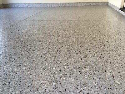 A close-up view of a speckled gray epoxy floor with subtle hints of white, black, and brown flakes, creating a terrazzo-like appearance. The light reflects gently on the surface, indicating a smooth and shiny finish. The background reveals a portion of a white wall meeting the floor at the bottom edge of the image.
