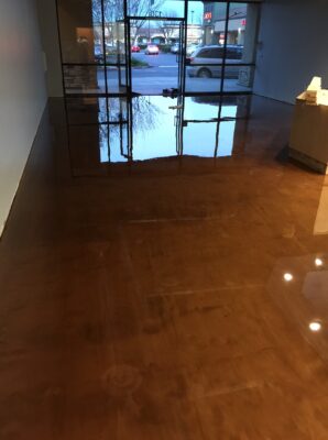 Polished warm-toned epoxy floor inside a commercial space with large glass windows, reflecting the evening sky and parking lot outside.