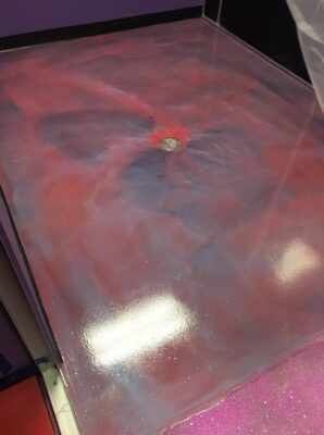 A vibrant epoxy floor with a swirling mix of pink, purple, and red hues, accented with white and shimmering specks to create a cosmic nebula effect. A drain is centrally located, around which the colors converge, resembling a spiral galaxy. The floor's glossy finish reflects the light above, enhancing the depth and dynamism of the colors. This flooring is bordered by purple and black walls, suggesting a bold, creative interior space.