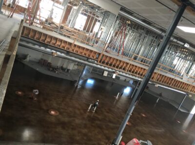 Elevated view of a construction site inside a large building with exposed structural elements. The floor below has a polished brown finish and scattered construction materials. Metal studs and beams reveal the framework of future walls and ceilings. Wooden joists support an upper level where more construction is underway. The environment is well-lit with natural light, and the site appears to be in the early stages of interior development.