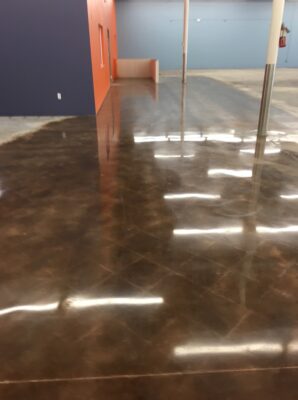 View of a polished brown concrete floor in a large room, reflecting fluorescent ceiling lights. The room features a dark blue wall on the left and a bright orange accent wall straight ahead with a bench and fire extinguisher. Metal support poles are evenly spaced throughout the space. The high gloss finish of the floor gives the room a bright and clean appearance, and there is no visible furniture or occupant.