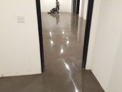 A view from a doorway into a brightly lit room with polished concrete flooring, reflecting light. On the far end, a person operates a floor cleaning machine, indicating ongoing maintenance. The walls are white, and the space is empty except for the maintenance activity. A light switch is visible on the wall to the left, highlighting the functional design of the space, which appears to be commercial or institutional.