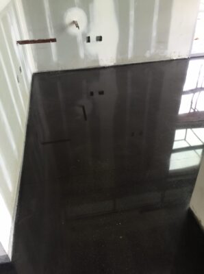 Image of a dark glossy concrete floor in a room with a white wall featuring electrical outlets and a metal push bar on a door. The reflection on the floor gives an illusion of depth, and the room appears to be empty and in the midst of construction or renovation. Natural light coming from the door window casts reflections and highlights the shiny surface of the floor.