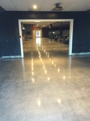 A hallway with polished concrete floors, leading into a well-lit room with a bar and stools visible in the distance. The hallway has dark blue walls and ceiling, with bright reflections from overhead lights casting a warm glow on the floor. The transition from the darker hallway to the inviting, brighter room ahead creates a welcoming atmosphere, likely in a commercial or hospitality setting.