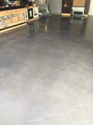 The image shows a section of a commercial space with a smooth grey concrete floor. On the left side, there is a wooden workbench with various items on top, and storage shelves filled with supplies. In the background, a tiled wall and a heater or air conditioning unit can be seen. The space gives the impression of a functional area, possibly a workshop or a back-of-house section in a retail store.