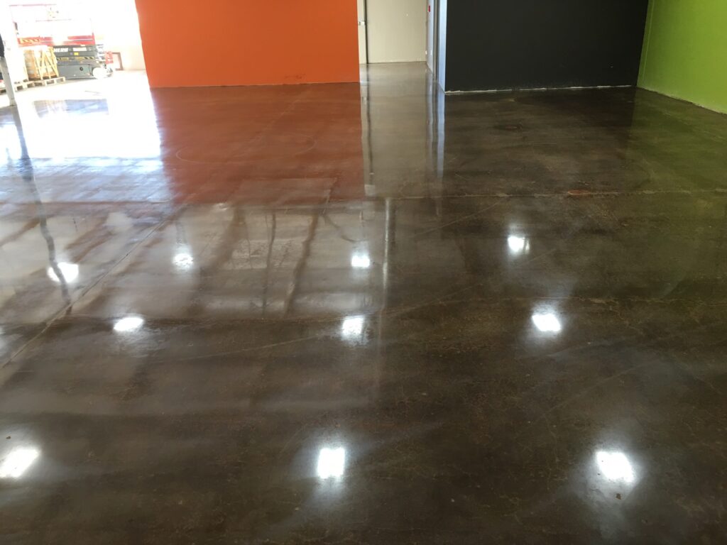 Image of a spacious room with polished concrete floors, reflecting the ceiling lights. The floor transitions from a warm brown to a darker shade towards the back. The walls are painted in bright orange on the left and lime green on the right, meeting a dark gray wall at the back. The space is empty, suggesting a commercial or industrial setting that is clean and well-maintained, with ample lighting.