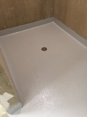 ALT Text: "A newly coated shower floor in a creamy white color with a textured, non-slip surface and a central round drain. The surrounding unfinished walls are lined with brown construction paper, indicating recent renovation or ongoing bathroom construction. The floor's slight incline towards the drain suggests proper grading for water flow.