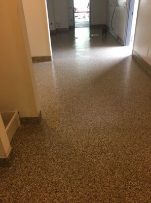 View of a hallway with a terrazzo floor consisting of small, multicolored specks on a tan background, giving a granulated appearance. The hallway is flanked by cream walls and a white baseboard with a brown accent. The flooring reflects the overhead lighting, creating a bright, polished look. The area is devoid of furniture, indicating a public or commercial space, possibly before the setup of equipment or furniture.