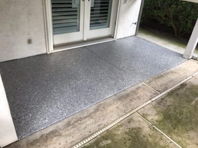 An outdoor patio area featuring a textured gray floor, edged by a white stucco wall and a single step leading to double doors with shutters. The floor is speckled with darker gray shades, suggesting a stone composite material. Surrounding the area are concrete slabs with visible moss growth, contrasting with the clean surface of the patio floor. An exterior electrical outlet is also visible on the wall to the left.