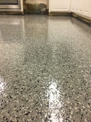 A glossy epoxy-coated floor with a speckled design of gray, white, and black chips creating a terrazzo-like effect. The sheen of the floor reflects the ambient light. The space suggests a residential setting, possibly a kitchen, with white cabinetry and a visible portion of a stove. There's evidence of wear or damage on the lower part of a cabinet, adding a lived-in feel to the scene.
