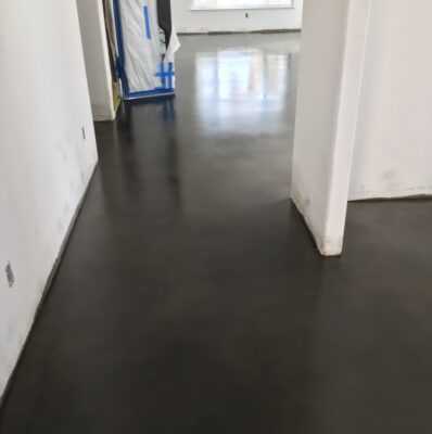 A hallway with a glossy polished concrete floor in a dark grey shade. The corridor is bordered by white walls with protective blue tape along the bottom, indicating a space under construction or renovation. The floor reflects the bright light from the open space ahead, creating a clean and modern industrial aesthetic.