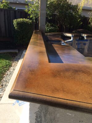 Angular perspective of a newly stained outdoor concrete countertop with a rich, warm brown tone and a smooth finish. The countertop is part of an outdoor kitchen or barbecue area in progress, as indicated by the protective blue tape and plastic sheeting in the background. The setting includes a garden with a wooden fence and shrubbery, suggesting a private residential backyard.