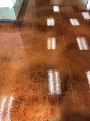 Close-up of a high-gloss stained concrete floor with a marbled effect combining rich tones of brown, red, and orange. The reflective surface showcases the detailed texture and variegation of the staining process, with overhead lighting adding bright spots to the polished finish. The edge of the floor is protected with blue tape, hinting at ongoing interior work or renovations.