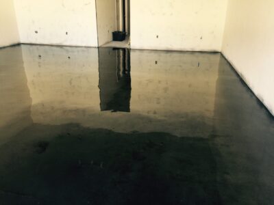 A corner of a room with a glossy concrete floor creating a mirror-like effect. The walls are white. Electrical outlets halfway up the wall are reflected in the concrete coating. A dark patch of water damage is visible on the floor, and a black trash bin is situated in the corner. The room is empty and the overall lighting is dim.