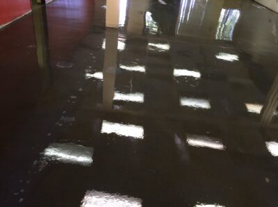 Glossy dark brown epoxy flooring in a commercial space reflecting overhead fluorescent lights. The surface is wet with scattered puddles, suggesting recent cleaning or a spill. Pillars and a bright red wall are partially visible, adding contrast to the scene. The reflection of the lights on the wet floor creates a pattern that enhances the depth and spaciousness of the area.