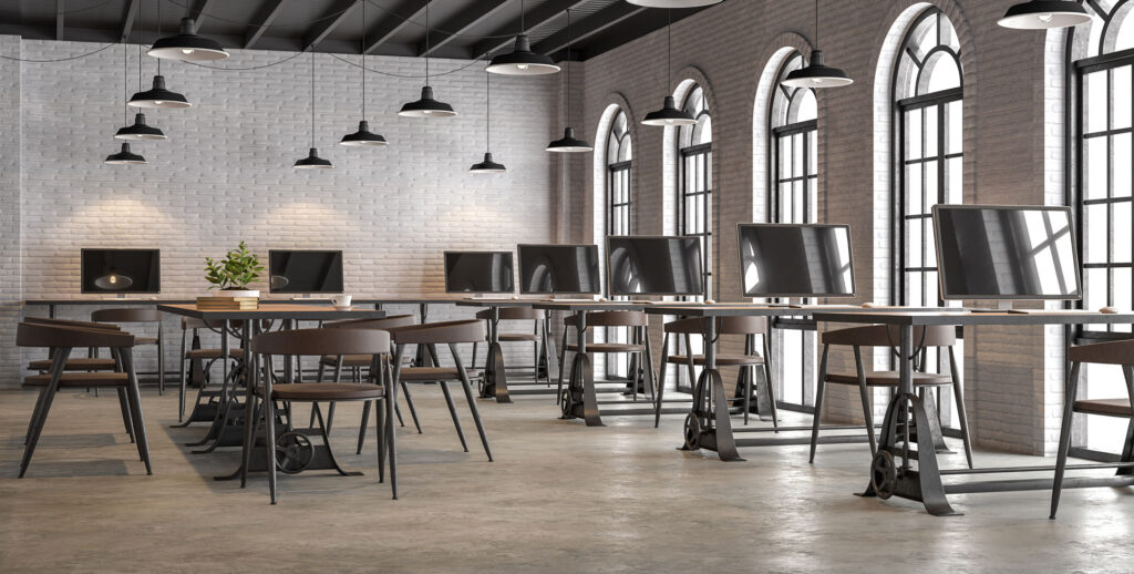 Spacious and well-lit industrial-style co-working space with high ceilings and large arched windows. The room features a series of wooden workstations with modern desktop computers, each paired with a black ergonomic chair. Overhead, black industrial pendant lights hang from the exposed beam ceiling, casting a warm glow on the white brick walls and the floors that have benefited from concrete polishing. The room is designed to foster a productive atmosphere with a blend of contemporary furnishings and urban architectural elements.