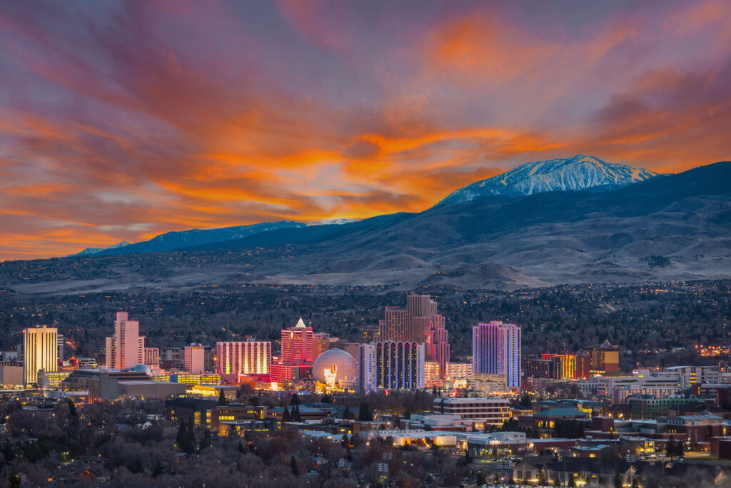 A picturesque view of a cityscape at dusk with a dramatic sunset sky. The horizon is adorned with vibrant shades of orange and pink clouds, contrasting with the snow-capped mountain in the background. The city is illuminated with the lights of high-rise buildings, creating a warm glow that reflects the urban life amidst the natural beauty of the surrounding landscape.