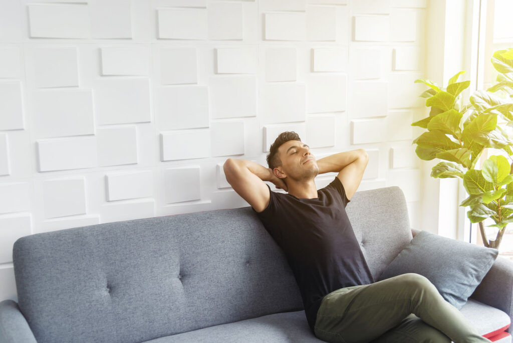 Relaxed man sitting on a grey sofa with his hands behind his head, enjoying a moment of relaxation in a bright room with a white textured wall and a green potted plant representing a light space that is allergen free.