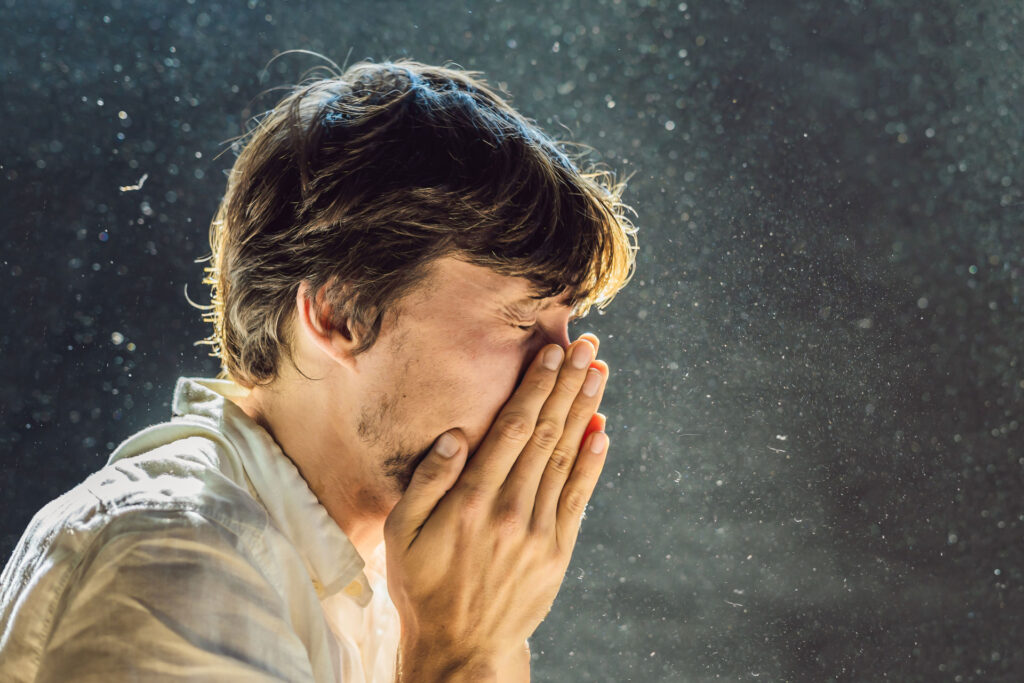 Close-up of a person sneezing with their hands covering their nose and mouth. The sneeze is captured mid-action, with droplets visible in the air against a dark background, highlighting the concept of spreading germs. The individual appears to have a concerned expression, emphasizing the importance of covering one's mouth while sneezing to prevent the spread of illness.