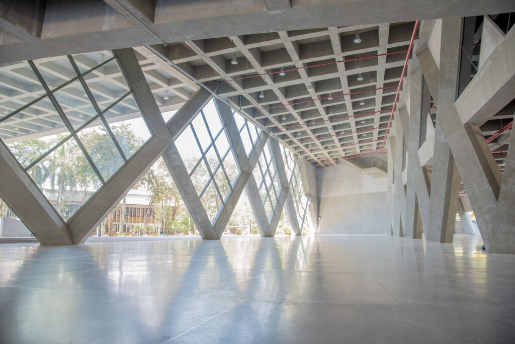 Interior view of a modern building featuring a geometric framework of concrete supports with large glass panels allowing natural light to filter in. The intricate pattern of the supports casts soft shadows on the polished concrete floor, creating a dynamic interplay of light and structure. The visible ceiling above showcases exposed beams and utilities painted in neutral tones with red lines, hinting at the industrial design of the space.