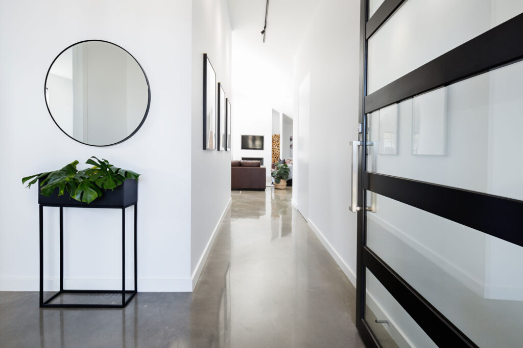 Elegant entryway in a modern home with a polished concrete floor, featuring a round mirror on the wall above a minimalist table with a potted plant, leading to a cozy living area.