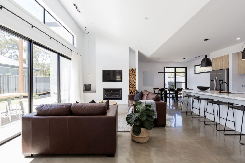 Modern open-plan living space with polished concrete floors with a gloss finish, featuring a kitchen with a breakfast bar, dining area, and a lounge with a brown leather couch, fireplace, and large windows allowing ample natural light.