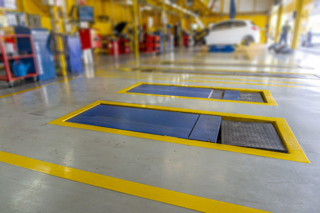 A close-up of the glossy epoxy floor of a vehicle service area with yellow safety markings around service pits, with blurred automotive equipment and vehicles in the background.
