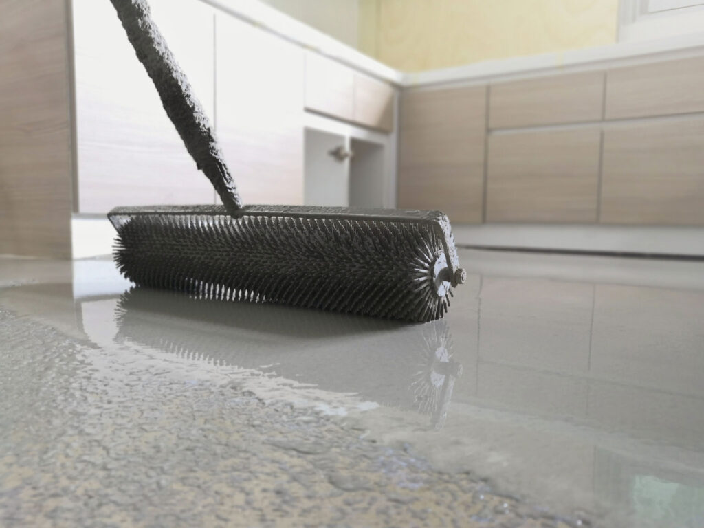 A paint roller with a black handle being used on a grey concrete floor, leaving a wet, shiny, and freshly painted surface, with a kitchen cabinet in the soft-focused background.